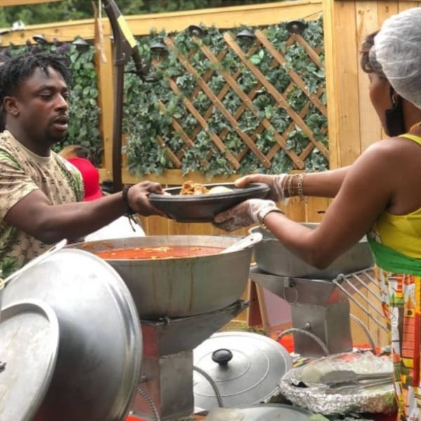 A man serving African food from a large pot to a woman at an outdoor event.