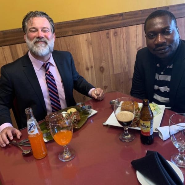 Two men seated at a table with African dishes and drinks at Drelyse African Restaurant.