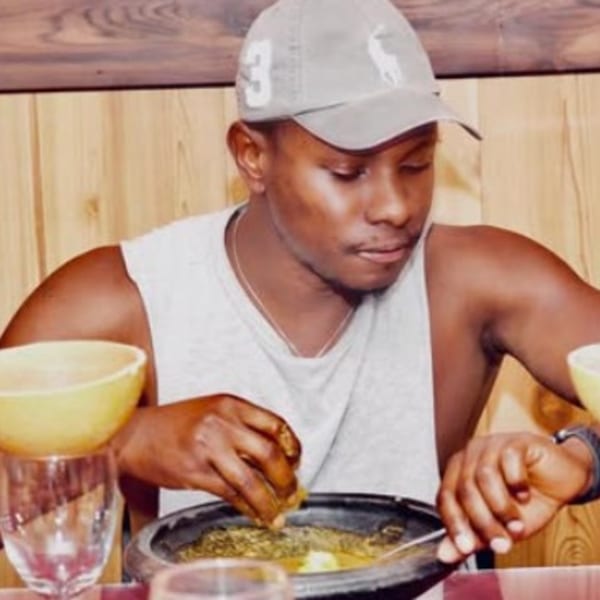 A man in a grey cap and tank top eats African food from a dark bowl with his hands.