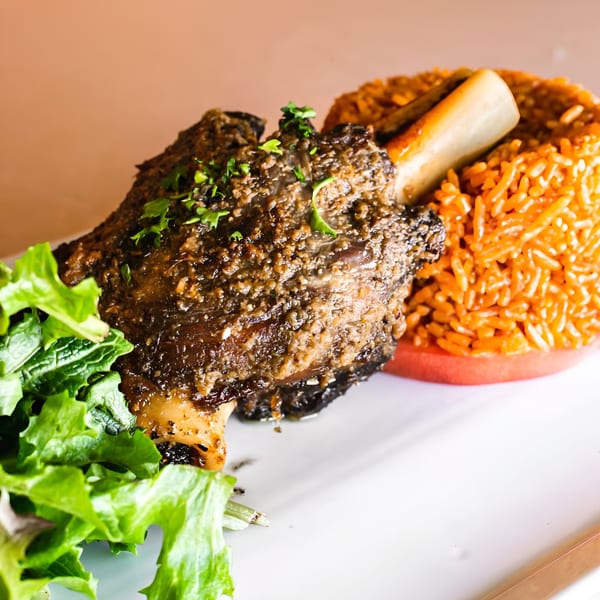 Braised lamb shank with jollof rice and fresh green lettuce on a white plate.