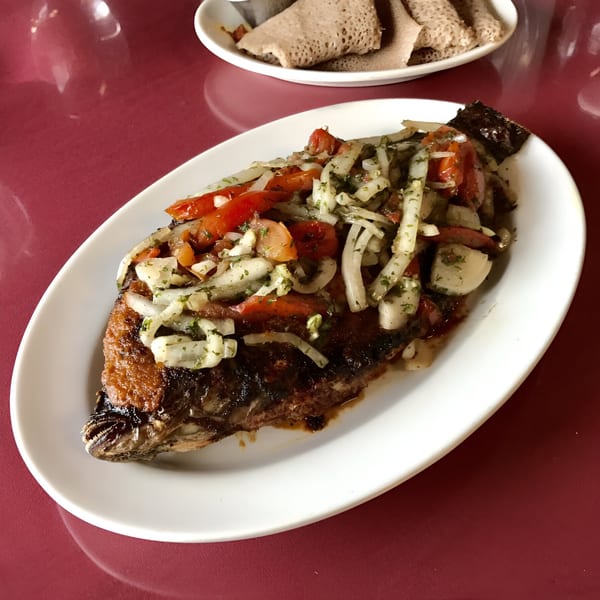 Whole fried fish topped with diced tomatoes and onions, served on a white platter with injera.