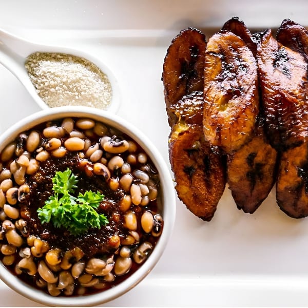 A bowl of African black-eyed peas and sauce with parsley, alongside fried plantains and a spoon of spice.