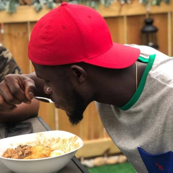 A man in a red cap and grey shirt eating an African dish from a white bowl.